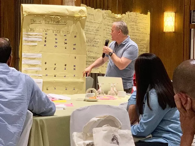 Roger Sweeney, a middle-aged, light skinned man with blonde hair stands in front of a seated audience and is pointing to a large notepad with writing on it. He is reviewing ex situ management in a workshop setting.
