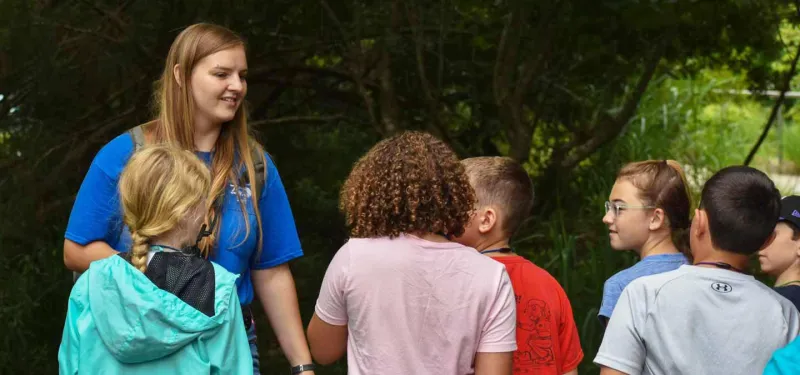 A female adult in a blue shirt with a logo is standing with a group of children outdoors. She is smiling and appears to be speaking to the group. The background is a mix of green foliage and trees.