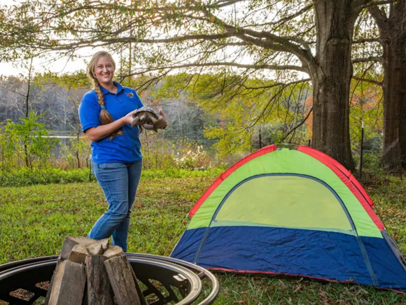 A woman with a long, blonde braid, wearing a blue collared shirt is standing next to a tent at a campsite in the woods. She is smiling at the camera and holding a very long, black snake in both hands.