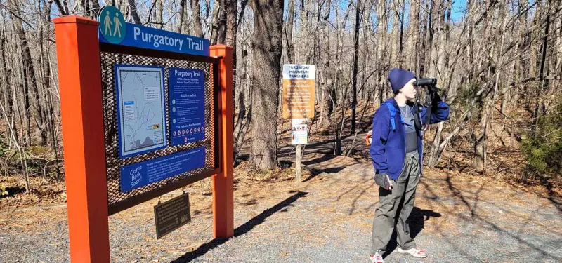 A person wearing a dark blue toboggan and jacket, stands at what appears to be a trail head in a forest, looking through a pair of binoculars. A large sign with the title "Purgatory Trail" appears to hold a map, and trail rules.