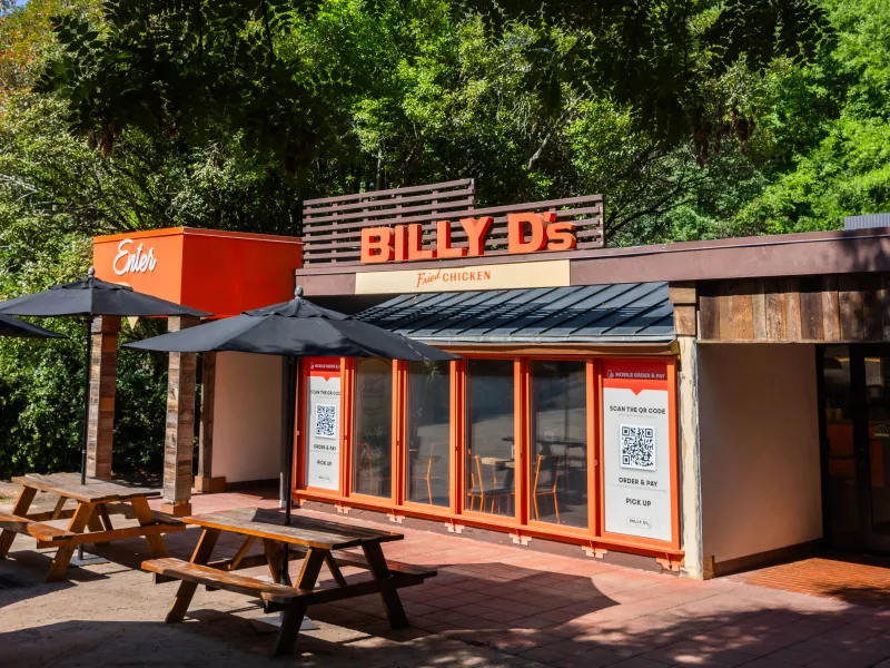A small restaurant named "Billy D's" with signage for fried chicken is situated amidst lush greenery. There are several outdoor picnic tables with black umbrellas on a paved area, providing shaded seating under the trees. The building features a modern facade with orange accents and QR codes displayed near the entrance for ordering.