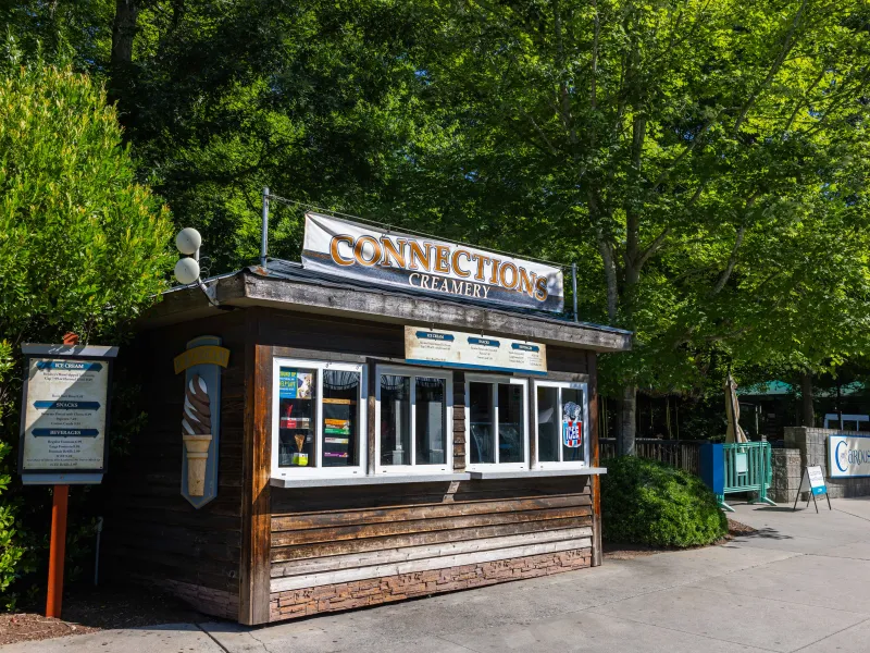 A small wooden ice cream stand named "Connections Creamery" with a vintage-style design. The stand features a large sign on top, windows for ordering, and a menu listing ice cream, snacks, and beverages on the side. It's surrounded by lush green trees on a sunny day.