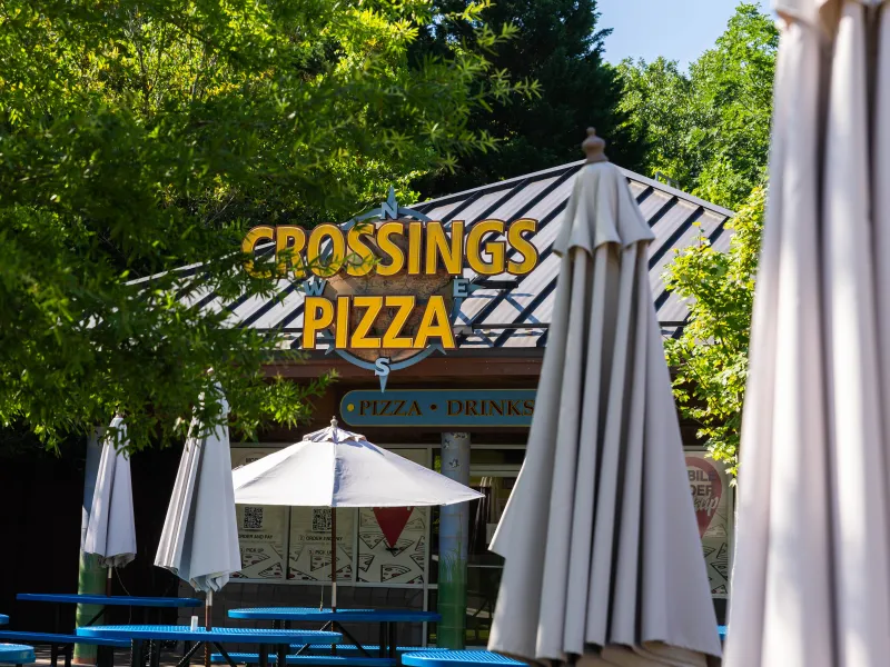 A pizzeria named "Crossings Pizza" with a metal roof is surrounded by lush green trees under a clear blue sky. A blue picnic table with a white umbrella is in the outdoor seating area, creating a welcoming atmosphere. Signs for mobile order pickup are visible on the restaurant's windows.