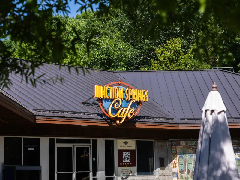 A cafe named "Junction Springs Cafe" with a dark roof and large yellow and blue sign. Green tree branches frame the top of the image, and a patio umbrella is visible on the left.
