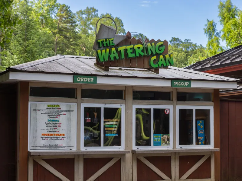 A small wooden snack kiosk named "The Watering Can" with a sign shaped like a watering can on top. It has two windows labeled "Order" and "Pickup." A menu is posted under the "Order" sign, offering snacks, drinks, and frozen treats. The stand is surrounded by lush greenery.