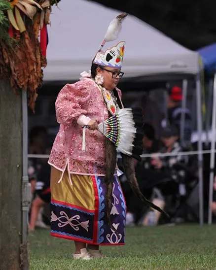 Native American woman wearing colorful traditional native clothing and holding a feathered and beaded fan.
