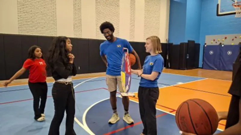 Two Zoo educators interact with youth at an indoor basketball court. The female educator holds a sign to allow a young girl to interact. The male educator holds a basketball while smiling broadly as the child interacts.