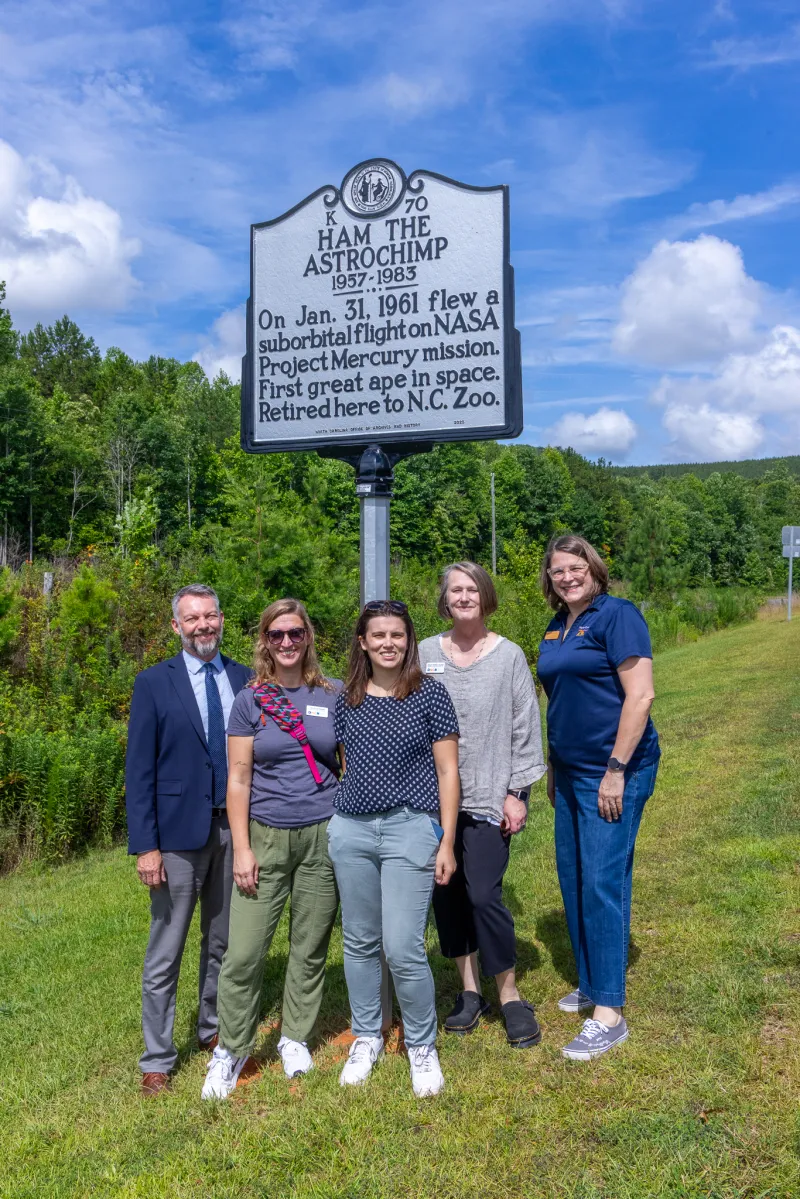 Members of Zoo leadership stand alongside DNCR staff beneath the official historical marker installed near the Zoo's main entrance; the marker is installed roadside in lush green grass, surrounded by thick forest and blue skies